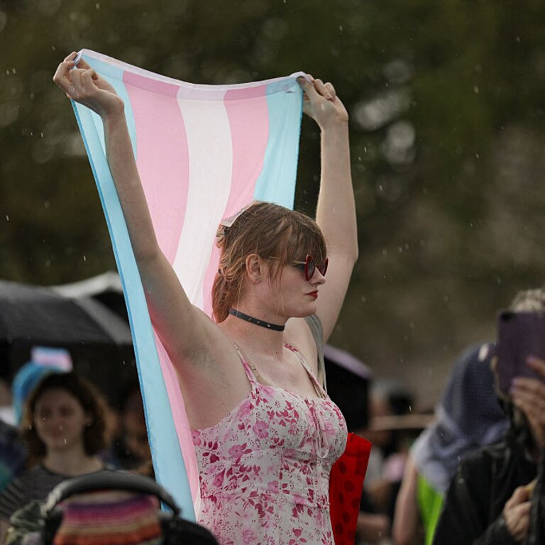 Eine Person steht in einem ärmellosen pink-weißen Top inmitten einer Menge im Regen und hält mit beiden Armen eine Trans-Pride Flagge über ihren Kopf | © Bryan Dozier | getty images