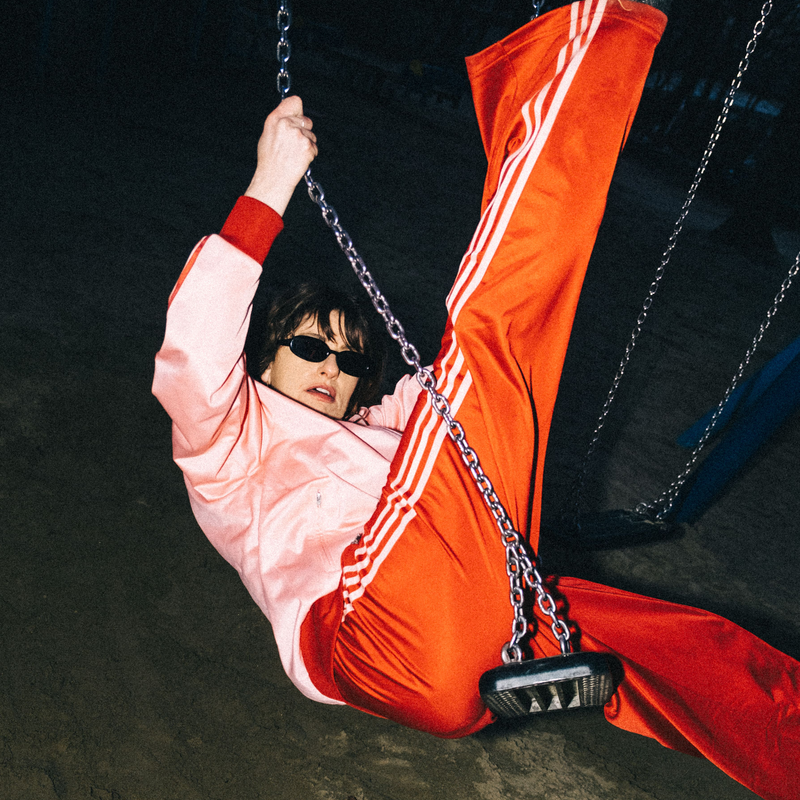 Die Künstlerin Mola sitzt auf einer Schaukel und trägt eine orangefarbene Sporthose sowie eine pinke Jacke. | © André Beiler