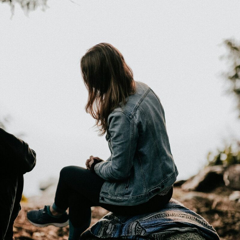 Frau sitzt am Wasser, Jeansjacke, lange braune Haare. Hintergrund grau. | © Foto: Priscilla du Preez | Unsplash