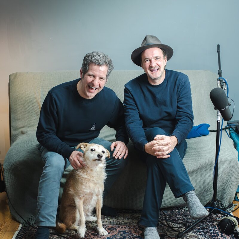 Die Podcast-Hosts Winson und Ueli sitzen im Aufnahme-Wohnzimmer, vor ihnen ein goldbrauner Hund, mittelgroß. Sie lachen in die Kamera. | © Foto: Winson