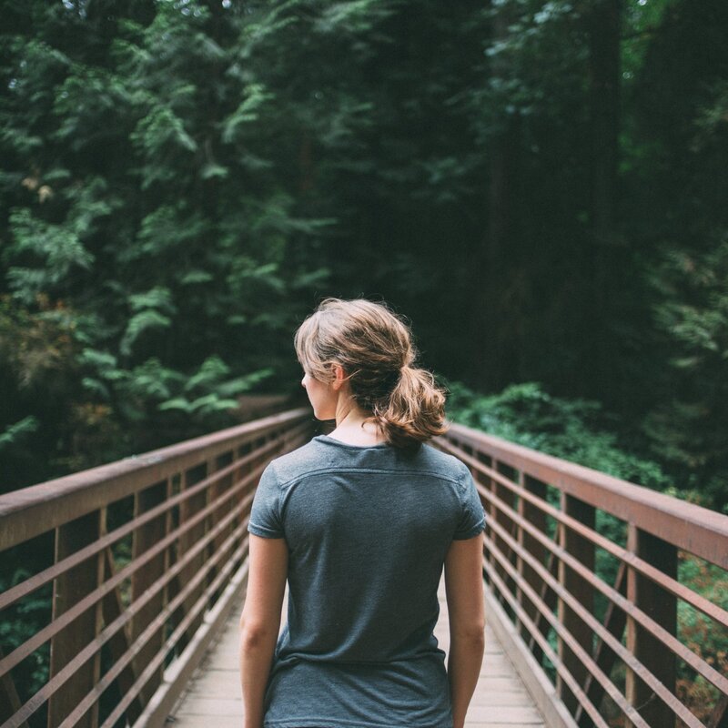 Eine Frau, von hinten zu sehen, geht über eine Brücke in Richtung Wald. | © Foto: Jake Melara/Unsplash