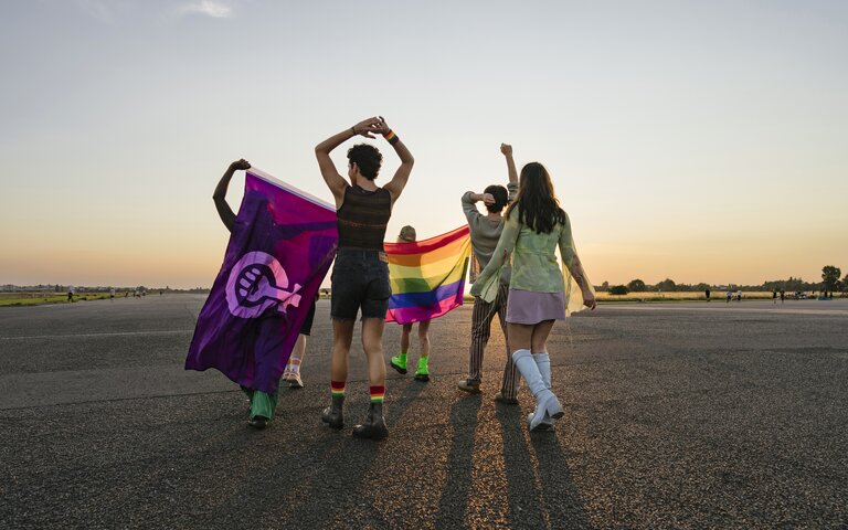 Vier Menschen mit dem Rücken zur Kamera gewandt vor einem Sonnenuntergang. Eine Person hält eine Regenbogenflagge und eine andere hält eine Feminismus-Flagge, | © Janina Steinmetz | gettyimages