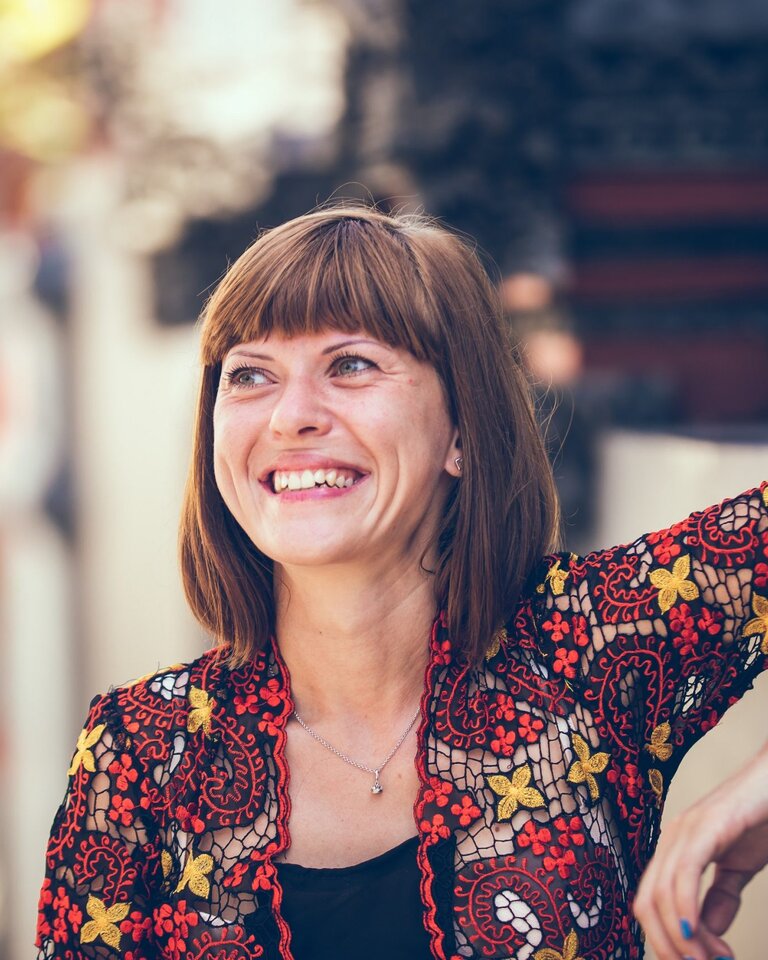 Frau mit halblangem Haar lächelt, Straßenszene. Sie trägt eine rote Bluse.