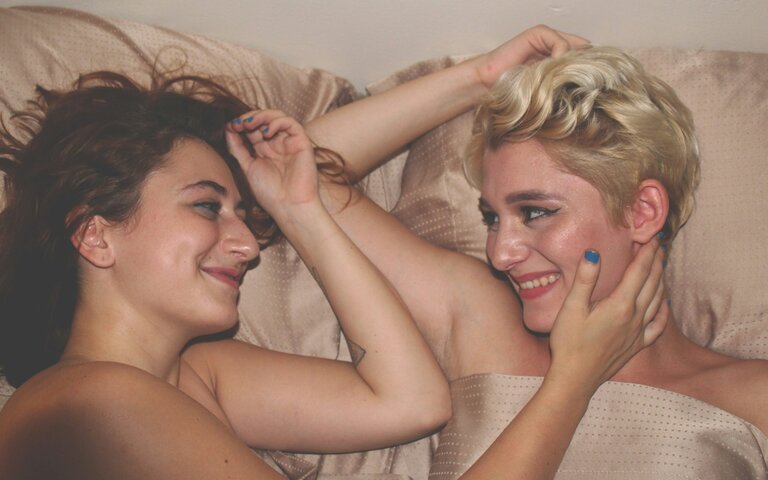 Zwei Frauen liegen nebeneinander im Bett, lächeln sich an. Sie sind nackt und berühren sich sanft mit den Armen. | © Foto: Mahrael Boutros | Unsplash