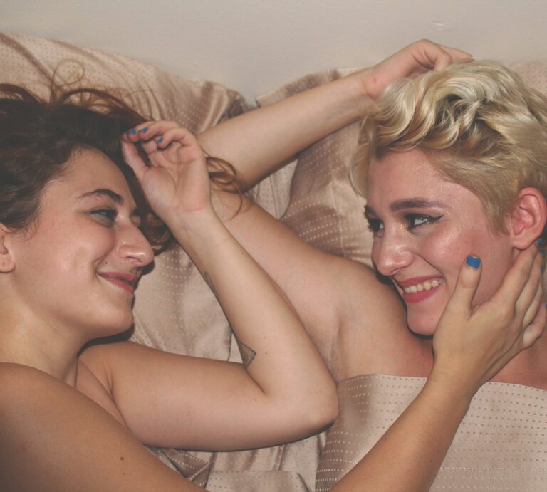 Zwei Frauen liegen nebeneinander im Bett, lächeln sich an. Sie sind nackt und berühren sich sanft mit den Armen. | © Foto: Mahrael Boutros | Unsplash