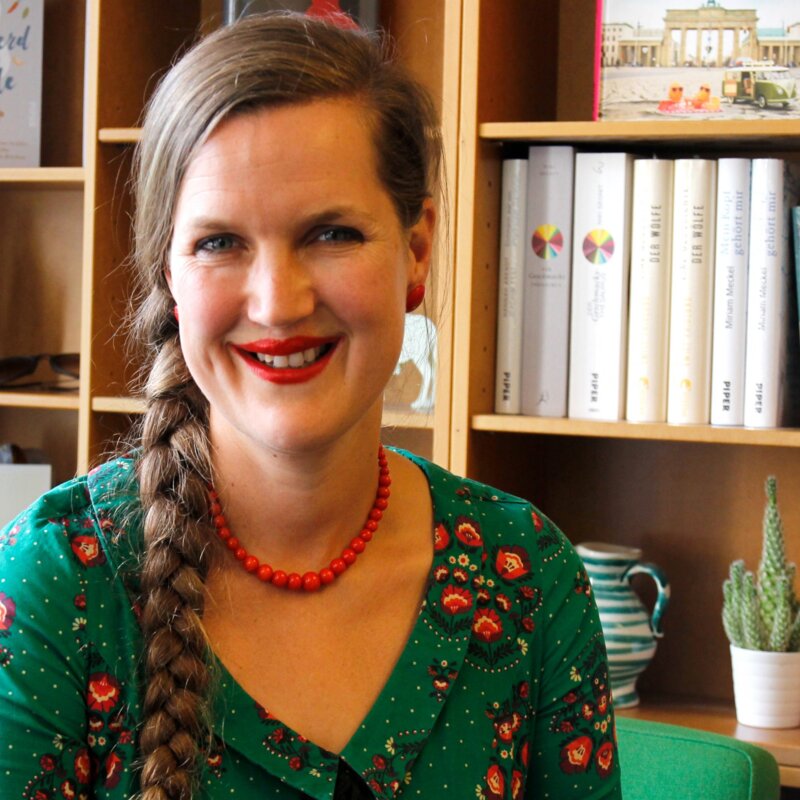 Frau in grünem Shirt blickt unmittelbar und lächelnd in die Kamera. Im Hintergrund steht ein Bücherregal mit vorwiegend weißen Buchrücken. | © Foto: Piper Verlag