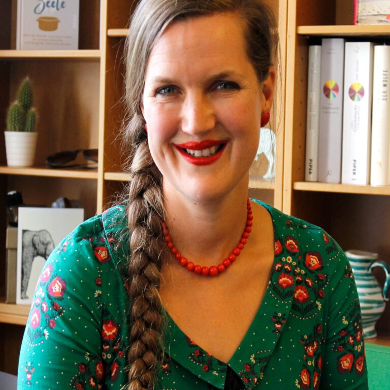 Frau in grünem Shirt blickt unmittelbar und lächelnd in die Kamera. Im Hintergrund steht ein Bücherregal mit vorwiegend weißen Buchrücken. | © Foto: Piper Verlag