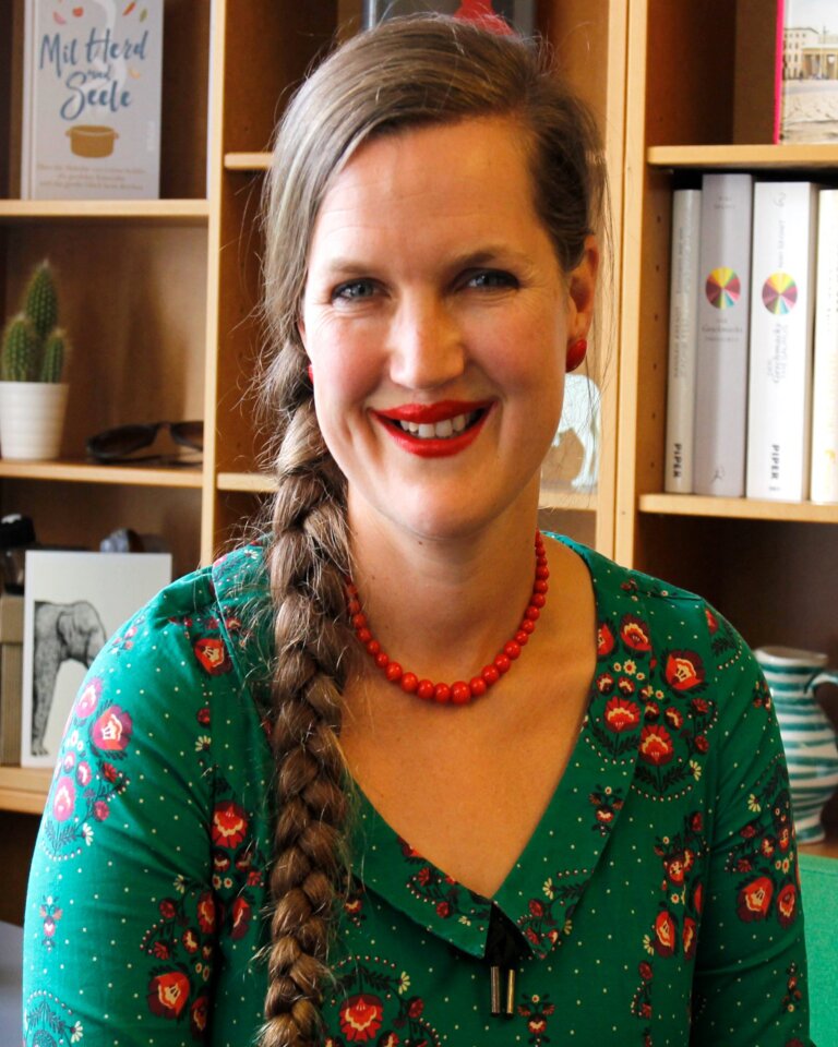 Frau in grünem Shirt blickt unmittelbar und lächelnd in die Kamera. Im Hintergrund steht ein Bücherregal mit vorwiegend weißen Buchrücken. | © Foto: Piper Verlag