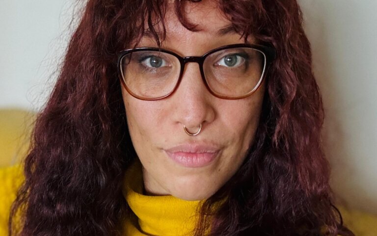 Das Foto zeigt Jasmin Dickerson. Sie hat lange rotbraune Haare, trägt eine Brille, einen gelben Pullover und sitzt auf einer gelben Couch. | © Die Autorin Jasmin Dickerson wünscht sich mehr Bewusstsein für Neurodiversität. Foto: privat