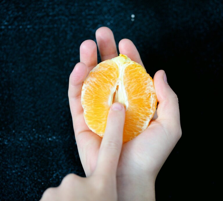 Eine Hand hält eine halbe, geschälte Orange, die eine Vulva symbolisieren soll. | © Foto: Sharon McCutcheon/Unsplash

