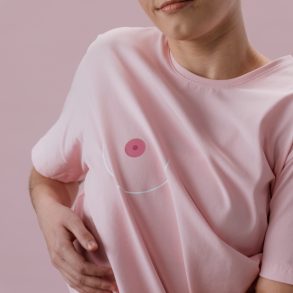 Zu sehen ist eine Frau in einem rosa T-Shirt, auf dem in Höhe der rechten Brustwarze das Bild einer Brustwarze aufgedruckt ist. Sie greift sich unter das T-Shirt.