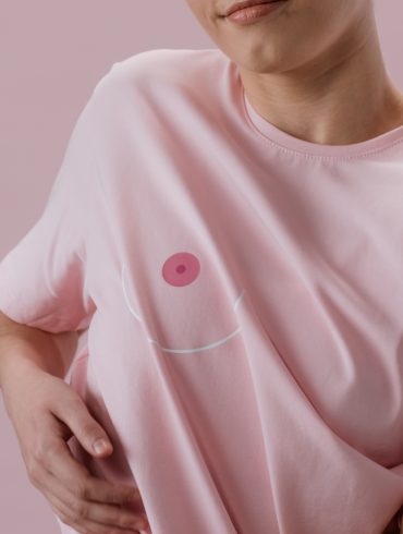 Zu sehen ist eine Frau in einem rosa T-Shirt, auf dem in Höhe der rechten Brustwarze das Bild einer Brustwarze aufgedruckt ist. Sie greift sich unter das T-Shirt.