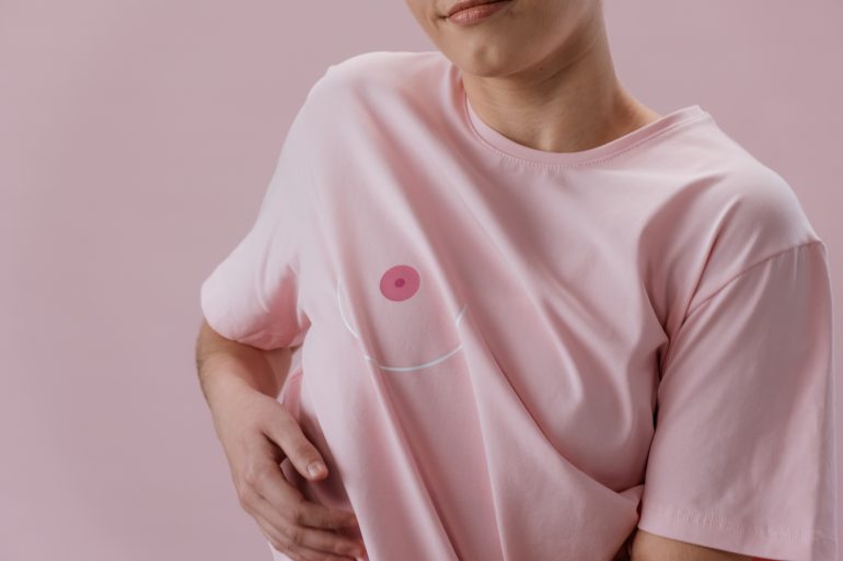 Zu sehen ist eine Frau in einem rosa T-Shirt, auf dem in Höhe der rechten Brustwarze das Bild einer Brustwarze aufgedruckt ist. Sie greift sich unter das T-Shirt.