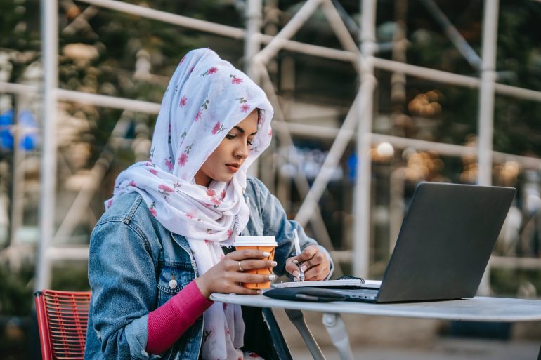 Eine Hijab tragende Frau sitzt mit einem Kaffeebecher in der Hand an einem Tisch und schaut in ihren Laptop.