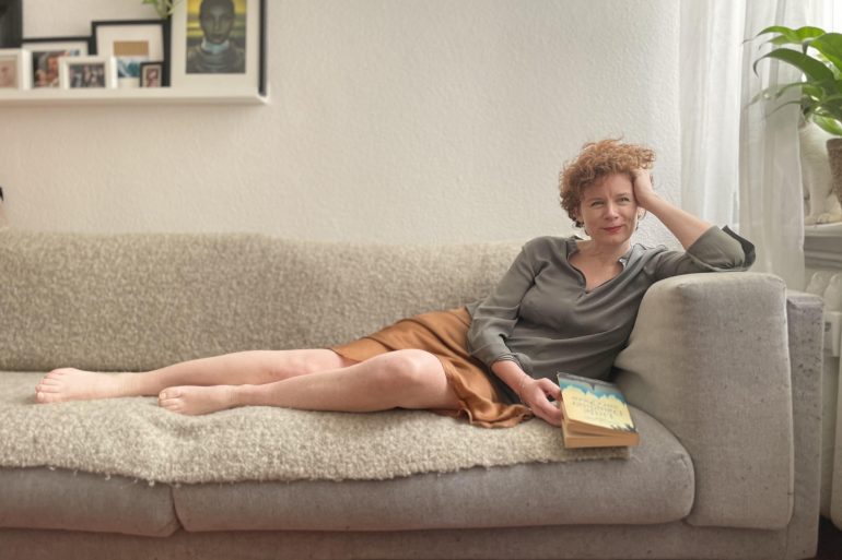 Auf dem Foto ist die Autorin Tina Molin zu sehen, die lächelnd und mit einem Buch in der Hand auf einem Sofa lehnt.