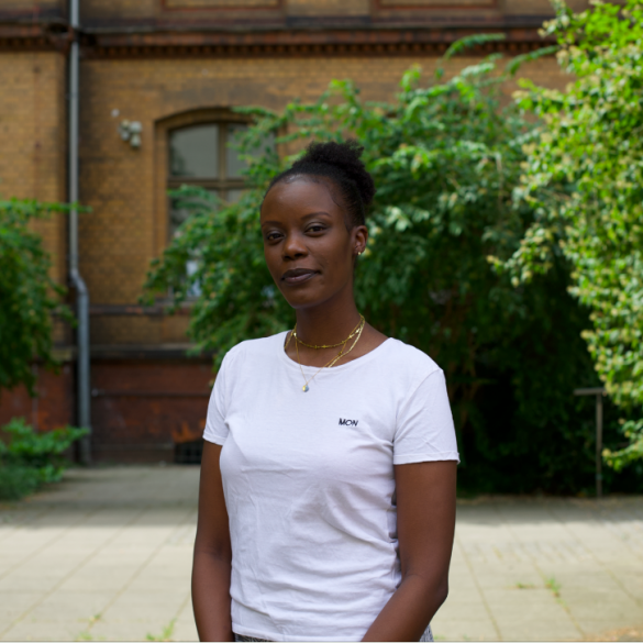Auf dem Foto ist die Autorin Anna Dushime in einem weißen T-Shirt zu sehen. Sie steht in einem grünen Innenhof und blickt in die Kamera.