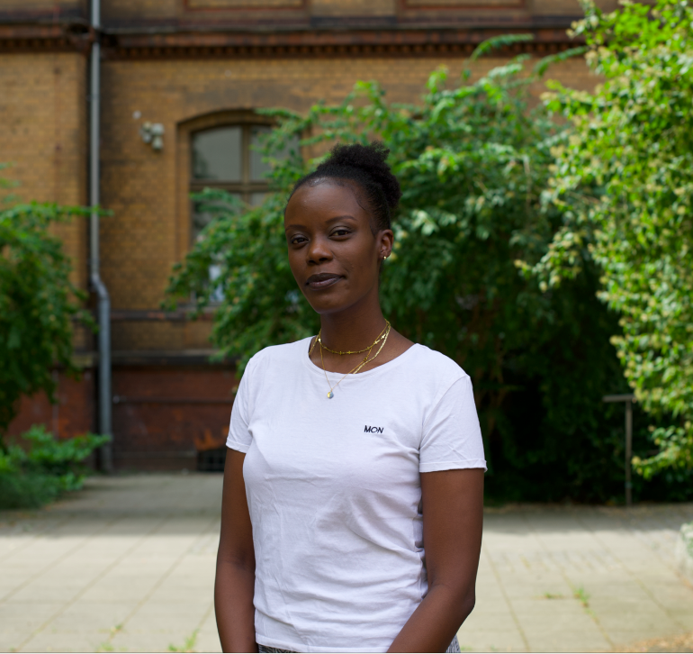 Auf dem Foto ist die Autorin Anna Dushime in einem weißen T-Shirt zu sehen. Sie steht in einem grünen Innenhof und blickt in die Kamera.