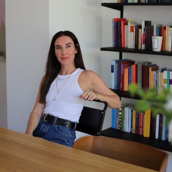 Auf dem Foto ist die Autorin Mirna Funk zu sehen, die an einem Holztisch sitzt und ernst in die Kamera blickt.
