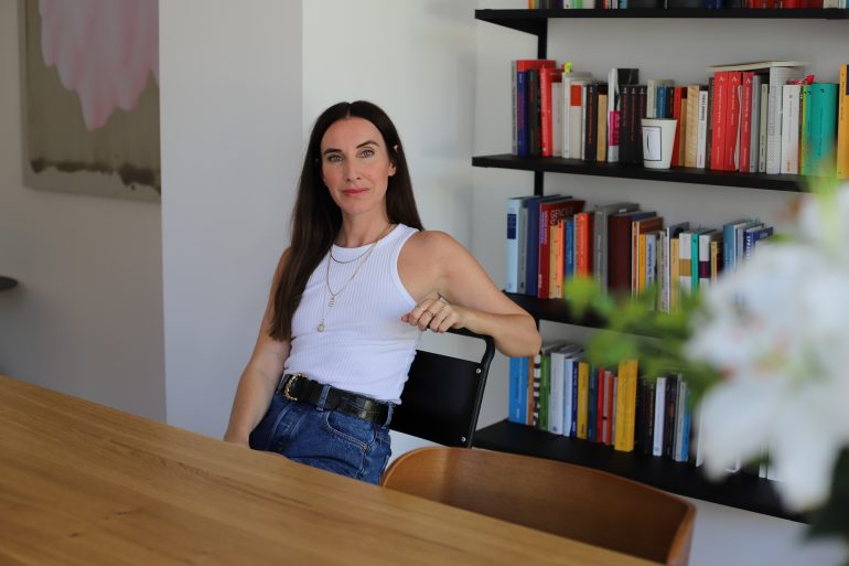 Auf dem Foto ist die Autorin Mirna Funk zu sehen, die an einem Holztisch sitzt und ernst in die Kamera blickt.