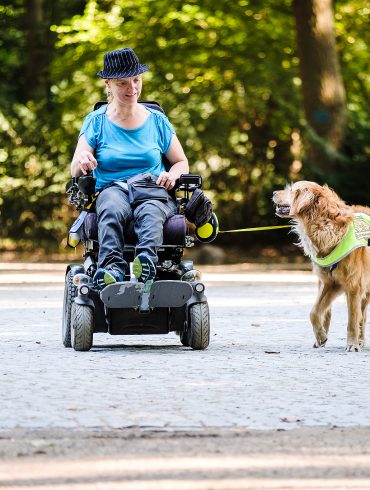 Eine Frau ist im Rollstuhl mit ihrem Assistenzhund im Park unterwegs.