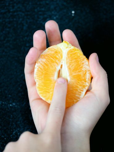 Eine Hand hält eine halbe, geschälte Orange, die eine Vulva symbolisieren soll.