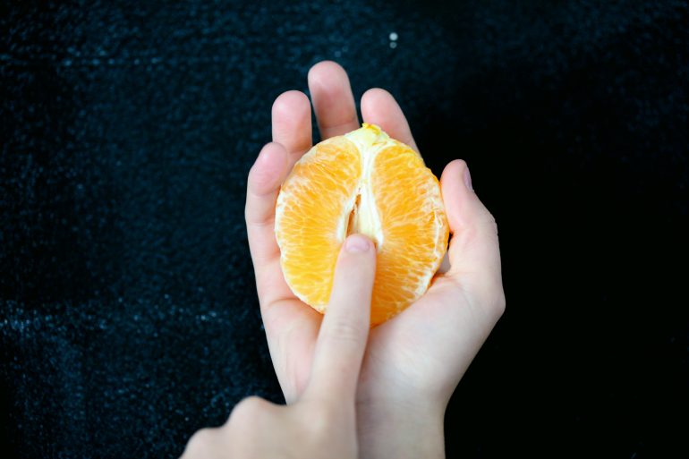 Eine Hand hält eine halbe, geschälte Orange, die eine Vulva symbolisieren soll.