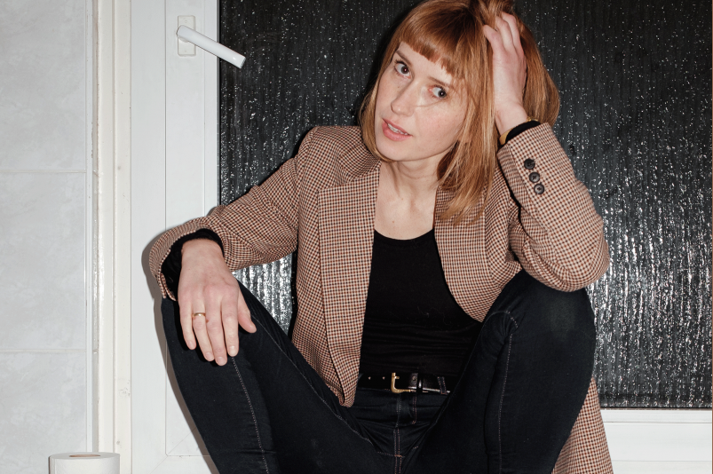Katja Lewina sitzt mit Schuhen und Jacke auf einer Heizung neben einer Klopapierrolle.