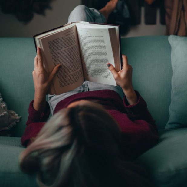 Auf dem Bild ist eine Frau von hinten zu sehen, die auf einem grünen Sofa liegt und ein Buch liest.