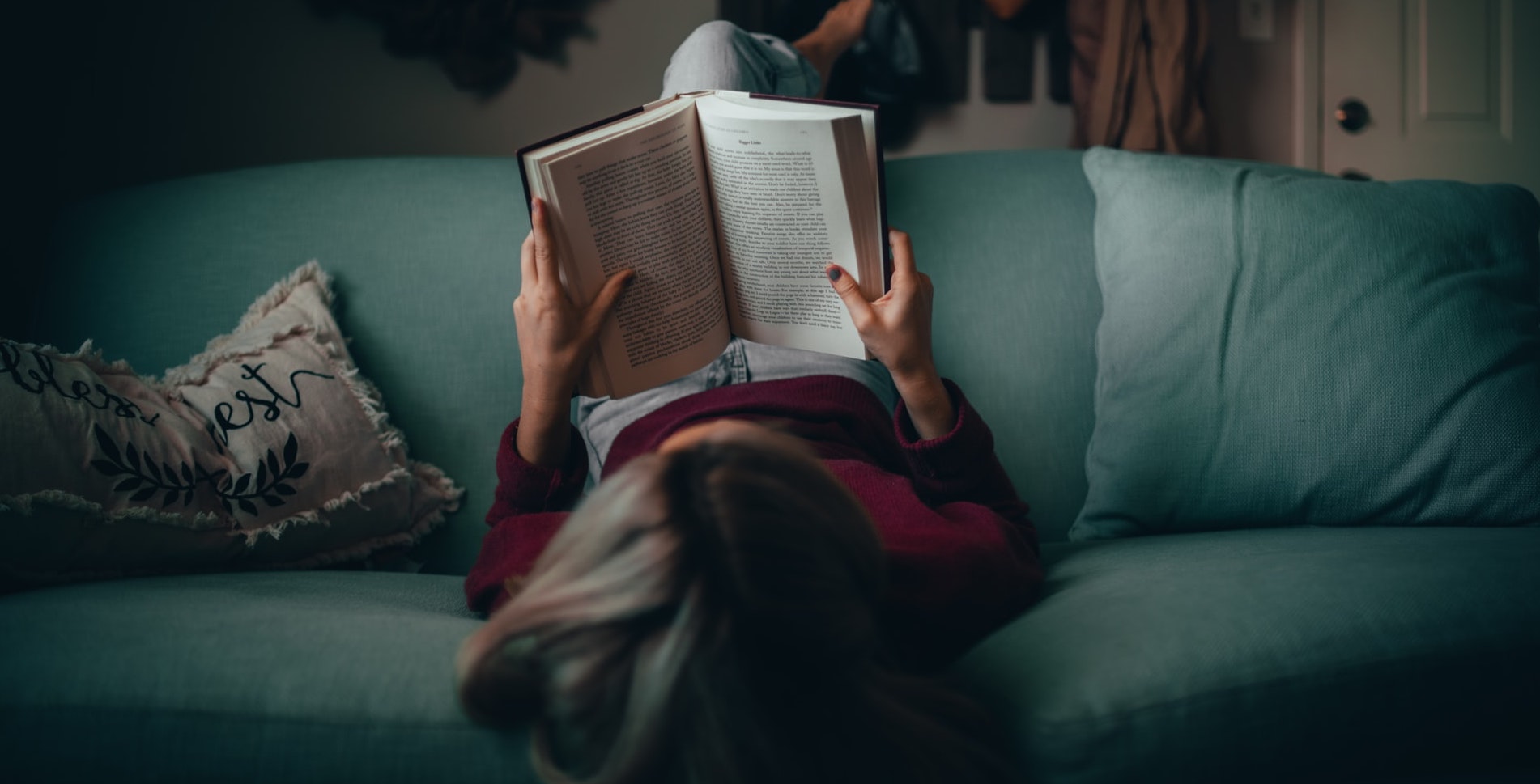 Auf dem Bild ist eine Frau von hinten zu sehen, die auf einem grünen Sofa liegt und ein Buch liest.