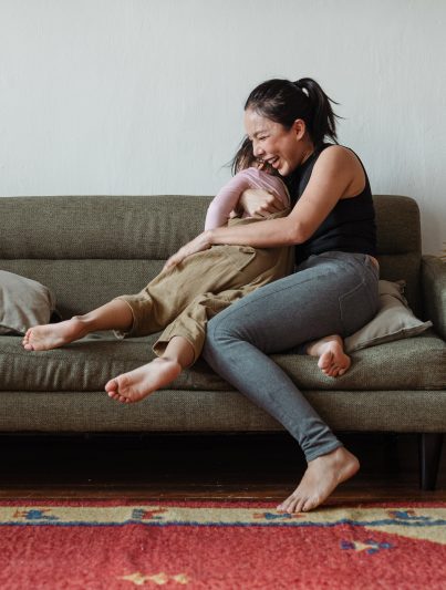 Auf dem Bild ist eine Frau mit dunklen Haaren und Jeans zu sehen, die auf dem Sofa liegt und ausgelassen lachend ihre Tochter in den Armen hält.