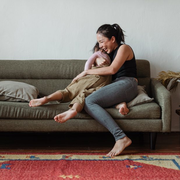 Auf dem Bild ist eine Frau mit dunklen Haaren und Jeans zu sehen, die auf dem Sofa liegt und ausgelassen lachend ihre Tochter in den Armen hält.