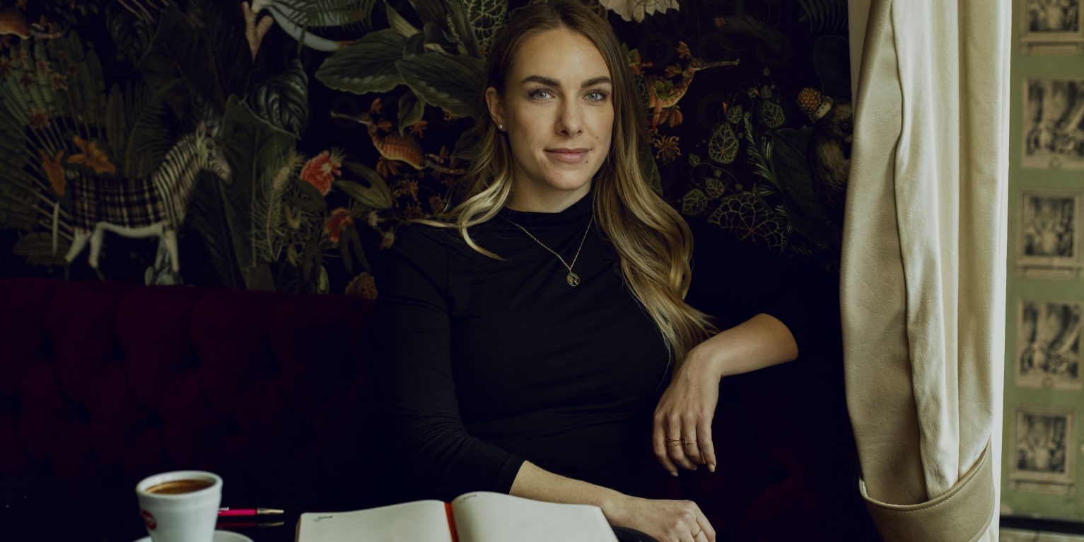 Protagonistin Gina Cohrs sitzt mit Kaffee und Buch in einem Café.
