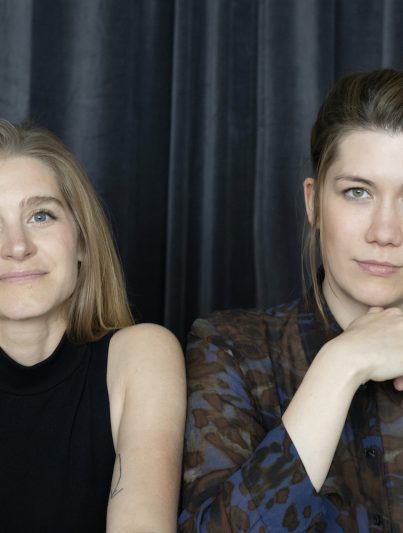 Die beiden Anwältinnen Katja Dunkel und Rebecca Richter stehen vor einem dunkelblauen Vorhang und blicken in die Kamera.