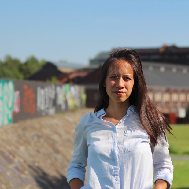Die Autorin Thuy-An Nguyen steht auf einer Freifläche mit einer besprühten Mauer und einer Wiese im Hintergrund, sie trägt eine weiße Bluse und blickt ernst in die Kamera.