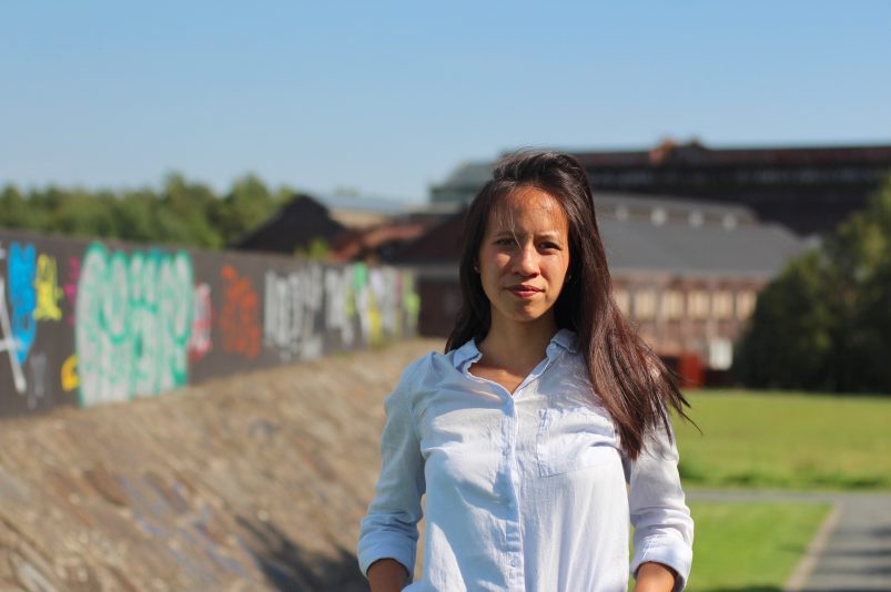 Die Autorin Thuy-An Nguyen steht auf einer Freifläche mit einer besprühten Mauer und einer Wiese im Hintergrund, sie trägt eine weiße Bluse und blickt ernst in die Kamera.