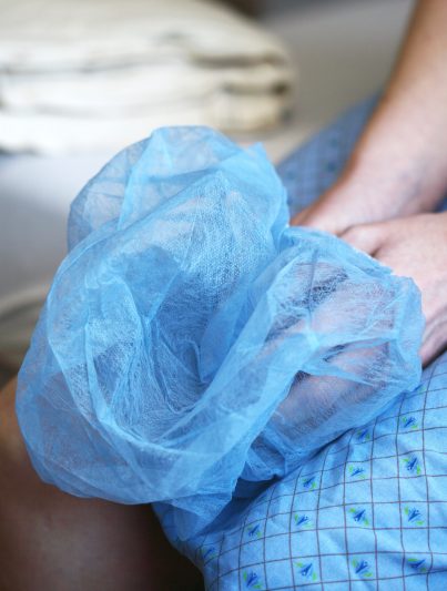 Auf dem Bild sind die Arme und Unterkörper einer Person zu sehen, die im Krankenhauskittel auf eine Operation wartet und dabei die OP-Kappe in den Händen hält.