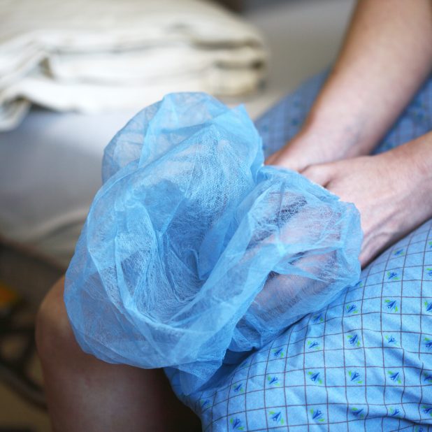 Auf dem Bild sind die Arme und Unterkörper einer Person zu sehen, die im Krankenhauskittel auf eine Operation wartet und dabei die OP-Kappe in den Händen hält.