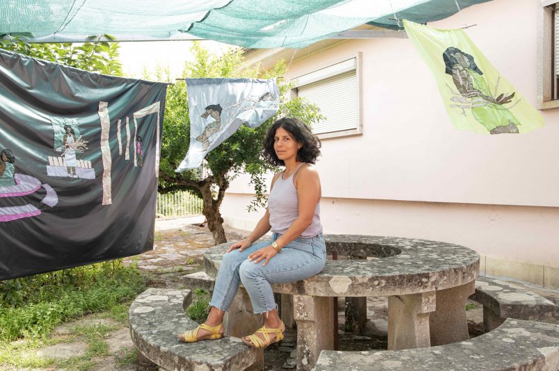 Die Künstlerin Sophie Utikal sitzt in einem Garten auf einer Steinmauer. Im Hintergrund wehen Stoffbahnen, die sie gestaltet hat.