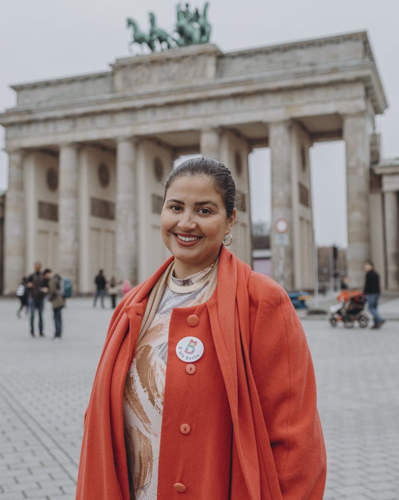 Alissa Hitzemann steht in einer roten Jacke und einem Sticker von „B wie Berlin“ vor dem Brandenburger Tor und lächelt in die Kamera.