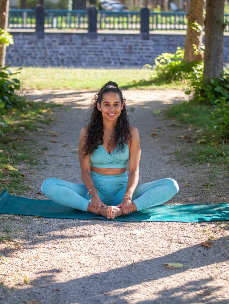 Die Coachin Mariela Georg sitzt auf einer blauen Yoga-Matte und lächelt in die Kamera. Sie ist in einem Park, mit grünen Bäumen und viel Sonnenschein.