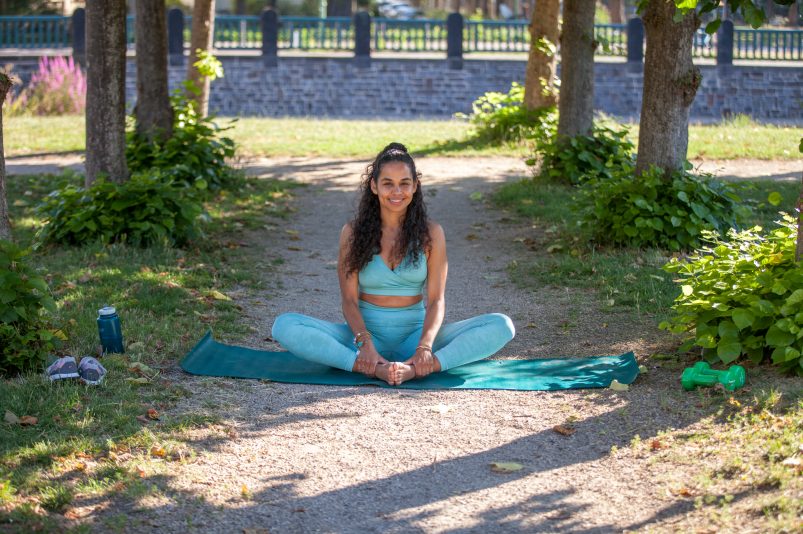 Die Coachin Mariela Georg sitzt auf einer blauen Yoga-Matte und lächelt in die Kamera. Sie ist in einem Park, mit grünen Bäumen und viel Sonnenschein.