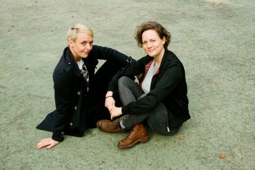 Susann Brückner und Caroline Kraft sitzen auf dem Asphalt. Sie tragen schwarze Hosen und Jackets.
