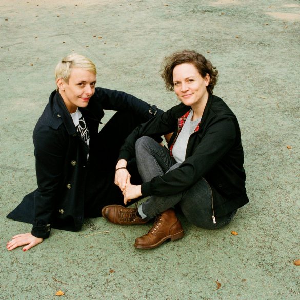 Susann Brückner und Caroline Kraft sitzen auf dem Asphalt. Sie tragen schwarze Hosen und Jackets.