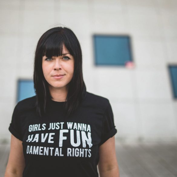 Mareike Fallwickl, Autorin des Buches „Die Wut, die bleibt“ steht im öffentlichen Raum vor einem Haus mit wenigen dunklen Spiegelfenstern. Auf ihrem T-Shirt steht der Schriftzug: Girls just wanna have fun. Demental Rights. :