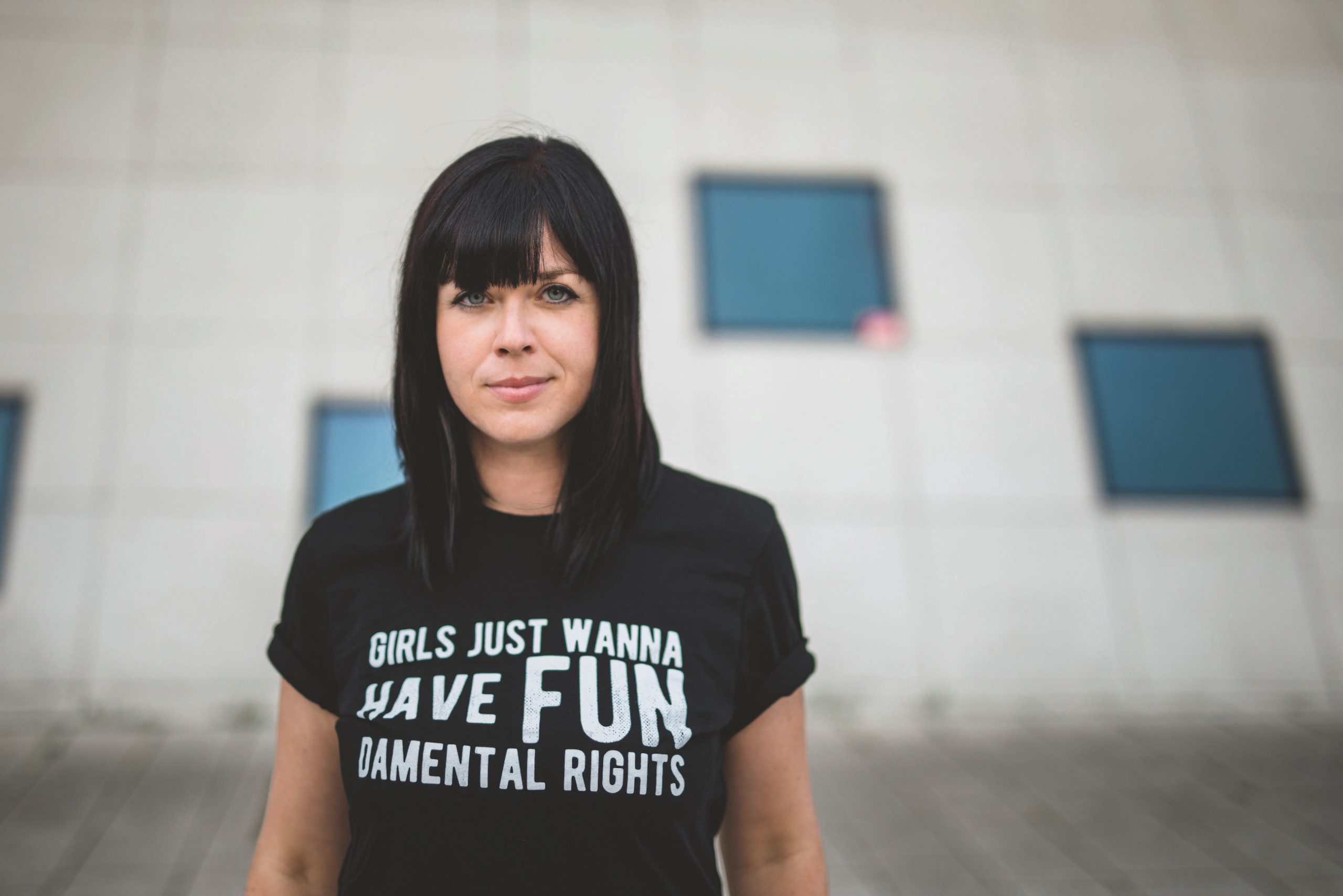 Mareike Fallwickl, Autorin des Buches „Die Wut, die bleibt“ steht im öffentlichen Raum vor einem Haus mit wenigen dunklen Spiegelfenstern. Auf ihrem T-Shirt steht der Schriftzug: Girls just wanna have fun. Demental Rights. :