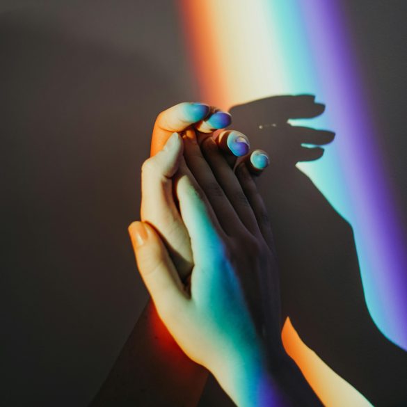 Das Bild zeigt zwei ineinander verschränkte Hände vor einer Wand. Die Wand wird mit einem Regenbogenstreifen beleuchtet.