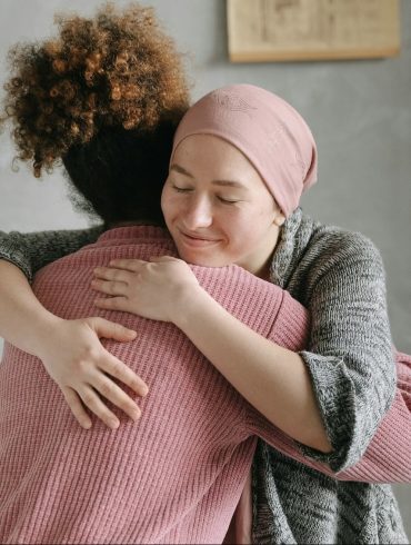 Zwei Frauen, eine davon mit einer aufgrund einer Krebserkrankung bedeckten Glatze, umarmen sich.