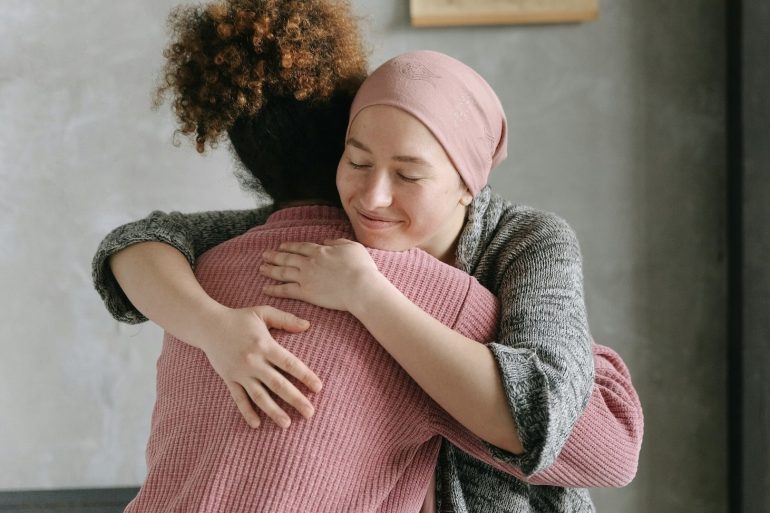 Zwei Frauen, eine davon mit einer aufgrund einer Krebserkrankung bedeckten Glatze, umarmen sich.