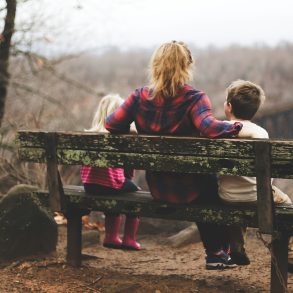 Eine Frau sitzt mit zwei kleinen Kindern auf einer Bank und schaut in die Natur.