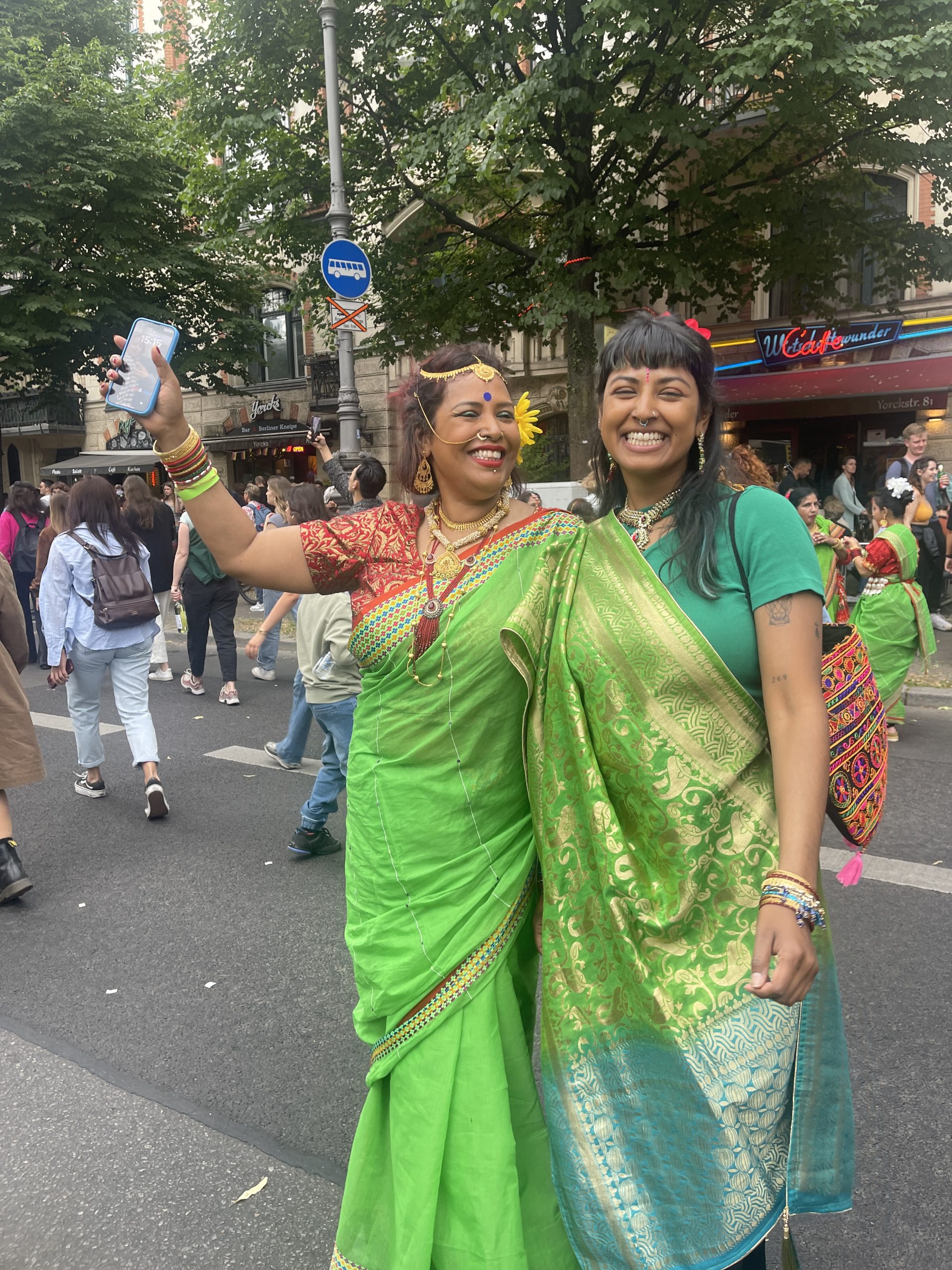 Das Foto zeigt zwei lächelnde Frauen auf einer Straße. Beide tragen ein grün-goldenes Tuch, einen Sari.